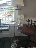 Two silver metal floor lamps side by side near a window, one with a white fabric shade, cords visible on floor.