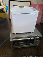 Side view of white Panasonic bread maker on top of silver Breville toaster oven on wooden floor.
