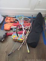 Full view of kids sports equipment including Nerf guns, lacrosse sticks, badminton racket, storage bag, and other toys on wooden floor.