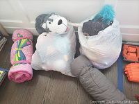 Two large white bags filled with assorted plush toys including a gray wolf plush, rolled-up pink sleeping bag with colorful designs, and gray sleeping bag bundled with penguin graphics on floor.
