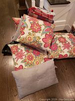 6 throw pillows, including 4 with bright floral pattern and 1 solid beige, stacked on hardwood floor with folded red and white linens on top.