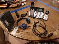 Full view of the lot showing AT&T cordless phone base and handsets, blue over-ear headphones, power cable, and Vonage router on wooden table.