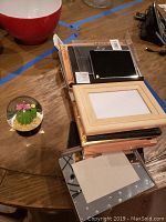 A collection of assorted picture frames stacked on a wooden table including wood, black, and silver frames; partially visible cactus snow globe on the side.