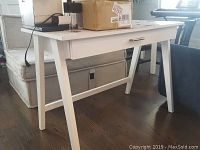 Full view of the white wooden desk showing the top surface, angled legs, and center drawer, placed on wooden flooring near sofa and other furniture.