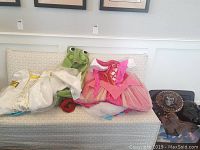 Photo showing multiple costumes including frog costume, princess dresses, and superhero outfit on couch