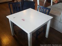 White square kids table with some surface stains and two blue sturdy wooden chairs around it on hardwood floor.