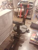 Side angle of the Gronk Fitness exercise machine showing seat, footrests, and red resistance straps attached to handlebars