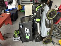 Front angle view of mower folded upright showing grass bag, battery charger with battery pack