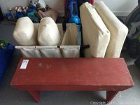 Wooden bench with reddish paint, showing wear marks, four ivory chair cushions, and Ikea cloth laundry sorter hamper with three compartments.