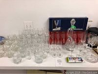 Full lot of assorted clear glassware including boxed Cristal D'arques wine glasses, decanters, various small and stemmed glasses, and barware accessories spread out on a table.