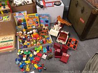 Wide-angle photo showing a collection of small children toys including colorful toy figures, vehicles, board books, and boxed art supplies, all arranged on a floor.