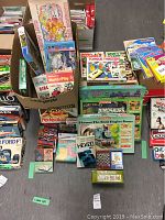Photo of assorted board games and puzzles stacked on floor, box art visible including Thomas the Tank Engine, Lost Valley Dinosaurs, Perfection, and other games.