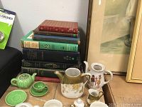 Photo of children's tea set items on a cardboard box in front of stacked antiquarian books and framed art
