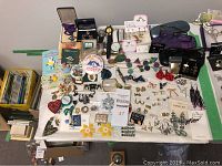 Full assortment of costume jewellery laid out on table including pins, earrings, brooches, watches, and glasses cases.