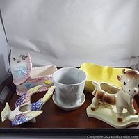 Lot of five vintage ceramic planters arranged on a wooden surface. Includes a pink and blue planter with ruffled edge, a white planter with matching saucer, a yellow rectangular planter, a calf-shaped planter, and a bird-shaped planter.