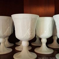 Close-up view of four milk glass goblets showing the raised grape cluster embossing on the bowl and plain pedestal base.