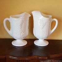 Front view of two white milk glass pitchers side by side showing grape cluster and leaf details with pedestal base.
