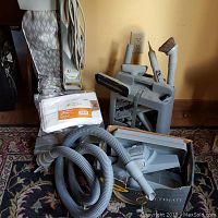 Kirby vacuum cleaner base unit with attached carpet shampooer, several gray plastic cleaning attachments, flexible vacuum hose, and power cord, all arranged on a carpeted floor against a wall.