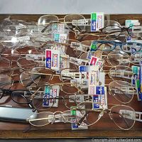 Top view of multiple pairs of reading glasses displayed on a flat surface with tags showing prices and magnification strengths. Various frame styles including aviator and rectangular in metal and plastic.