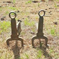 Pair of gunstock style metal andirons standing upright outdoors on dry grass and dirt, showing their front and side profiles.