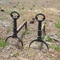 Pair of metal andirons set outdoors on the ground, showing overall structure and condition including the tripod feet and looped handles.