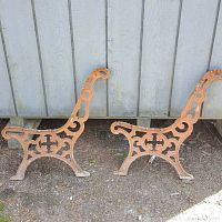 Side view of two cast iron park bench legs with intricate scrollwork and cross details. Legs show signs of rust and multiple paint layers.
