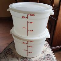 Two white plastic Cambro tubs stacked with visible red measurement markings on the sides showing the capacity graduated up to 12 quarts, each tub has a lid.