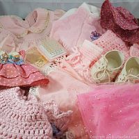 Assorted vintage pink baby doll clothes including knitted dresses, crochet booties, polka dot skirt, bonnet hat, and doll shoes