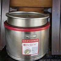 Front view showing stainless steel soup cooker with lid and red trim, placed inside a cabinet.