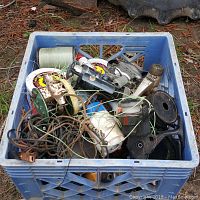 Blue plastic crate filled with various fishing reels, reel parts, fishing line spools, and fishing accessories outdoors on the ground.