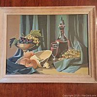 Front view of vintage paint-by-number still life painting showing bowl of grapes, wine decanter, glass, and fruit on draped fabric background, framed in light wood.