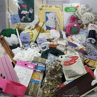 Box lot of assorted craft supplies including ribbons, cords, trims, pins, and embroidery sampler visible.