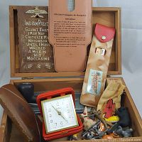 Open cedar dresser box showing assorted vintage items inside, including a folding square alarm clock, wooden plaque with Indian prayer, leather cases, and small figurines.