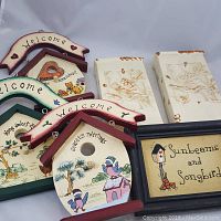 Photo of two cream ceramic wall pockets with embossed bird designs, three wooden hanging birdhouse decor pieces painted with colorful birds and floral patterns, and one framed fabric art piece with the words 'Sunbeams and Songbirds'.