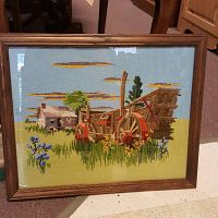 Framed crewel embroidery showing a wooden cart in a field with a sky background.