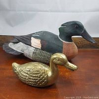 Wooden and brass duck figurines displayed on a wooden surface, showing size and detail comparison.