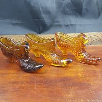 Three vintage yellow/amber art glass shoes displayed side by side on a wooden surface, showing detailed pressed glass patterns including hobnail and starburst motifs.