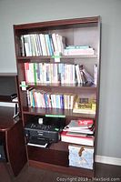 Front view of cherry finish bookcase showing four shelves with various books, printer, and files. Left side damage visible on the frame.