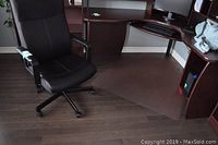 Office chair at a corner office desk with plastic floor protector under it on dark hardwood floor.