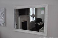 Framed rectangular wall mirror with ornate white frame hanging on a wall, reflecting part of a room including a fireplace and lamp.