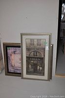 Two stacked framed prints leaning against wall, one with floral and the other showing a street scene