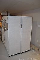 Two closet units side by side covered in light-colored fabric with black trim, showing their shape and positioning.