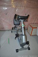 Side view of stationary exercise bike showing padded seat, backrest, and pedals with straps