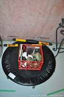Top view of small round trampoline with padded edge. Red crate containing weights and exercise accessories placed in the center. Yellow and black Cardio Blade exercise bar rests on trampoline edge.