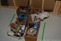 Photo showing five boxes of assorted household maintenance items arranged on floor, visible items include metal fittings, tools, and small parts