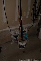 Photo showing open bags of ice salt and limestone screening behind a bucket of polymeric sand with three driveway brushes leaning against them in a garage corner.