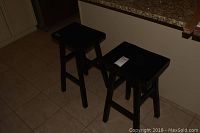 Two espresso colored wooden stools shown side by side on tile floor near granite countertop.
