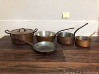 Photo of five copper pots and pans displayed on wood surface, showing varying sizes and handle types, all with visible tarnish and used condition.