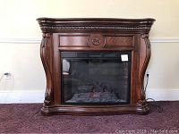 Front view of the large, solid wood electric fireplace showing detailed carvings, curved sides, and artificial log firebox behind glass.
