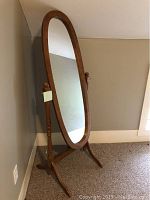 Solid maple framed long oval floor mirror with an adjustable wooden stand, shown standing against a wall on carpet flooring.